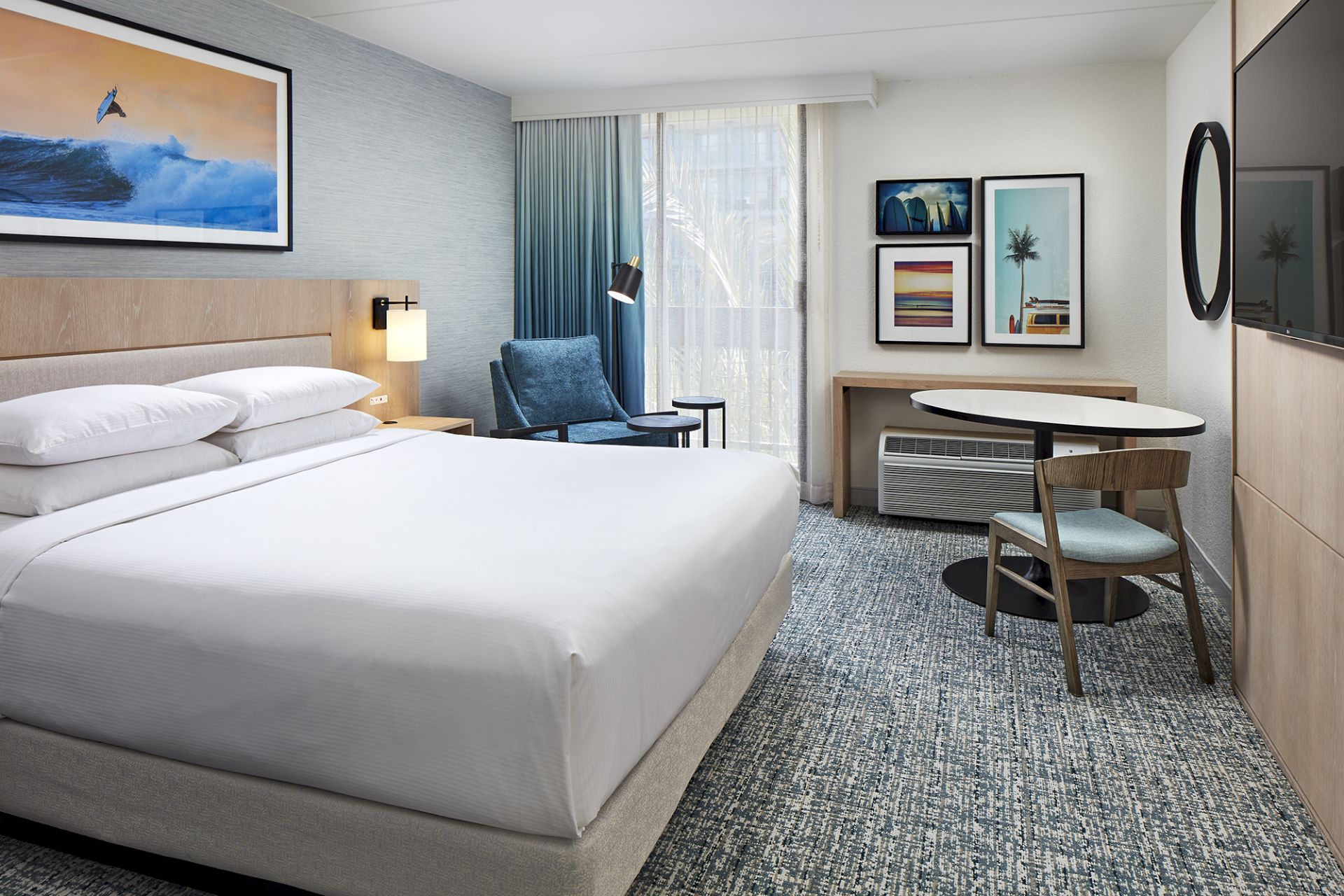 A modern hotel room with a large bed, blue curtains, seating, a desk and chair, artwork, and a window with light.