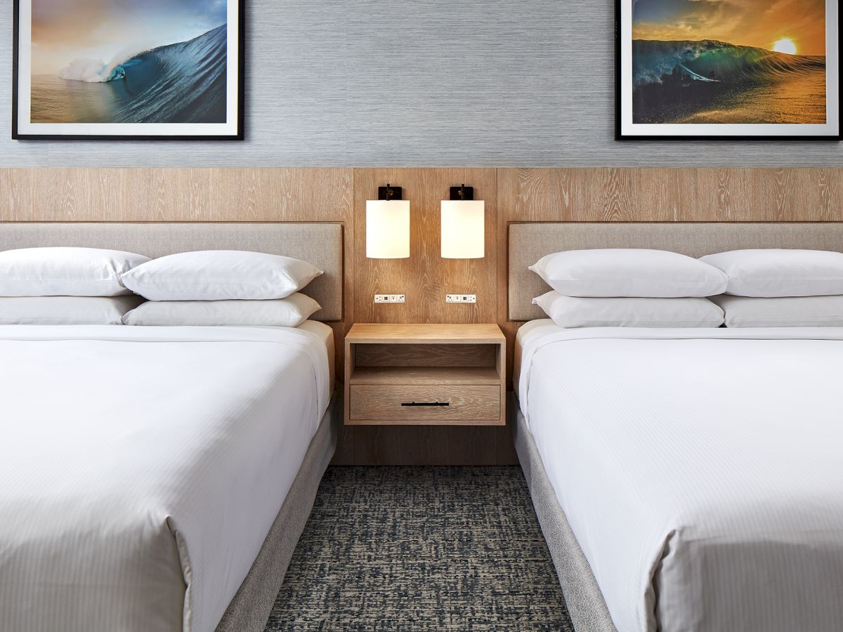 Two neatly made hotel beds with a shared nightstand, wall lamps, framed sea photos, and a patterned carpet.