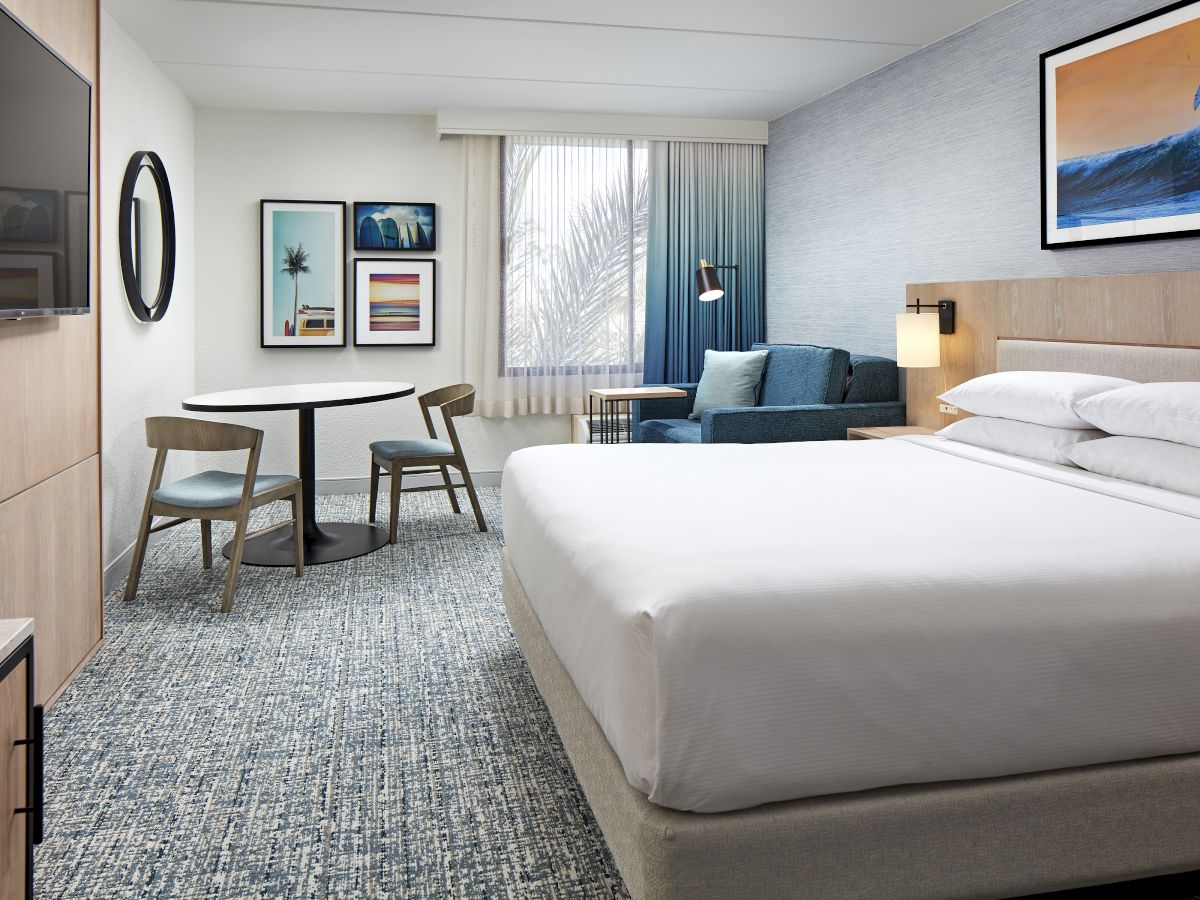 A modern hotel room with a large bed, seating area, desk, wall art, a window with blue curtains, and a patterned carpet.