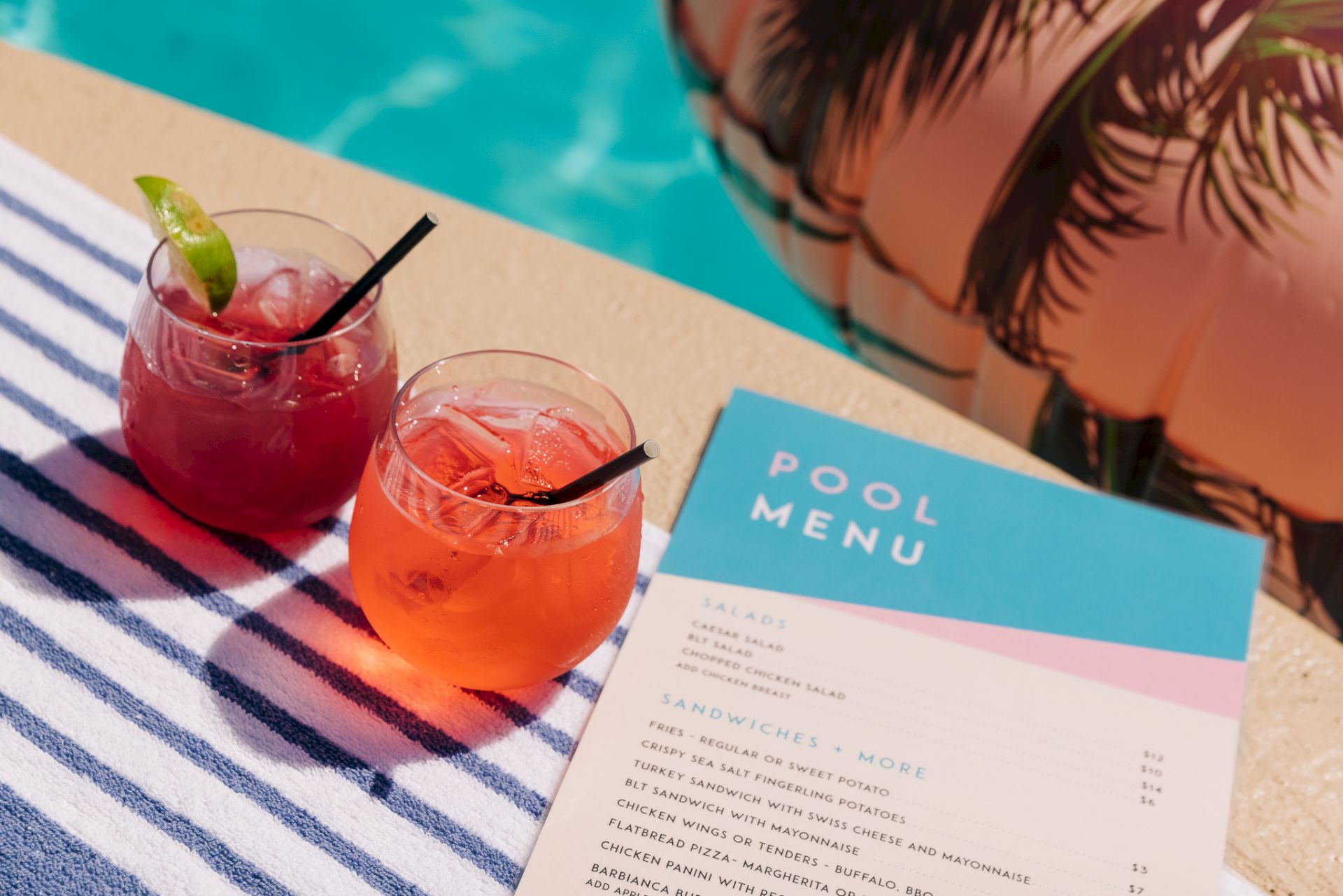 Two colorful drinks with ice and straws sit on a striped towel beside a pool, next to a pool menu card.