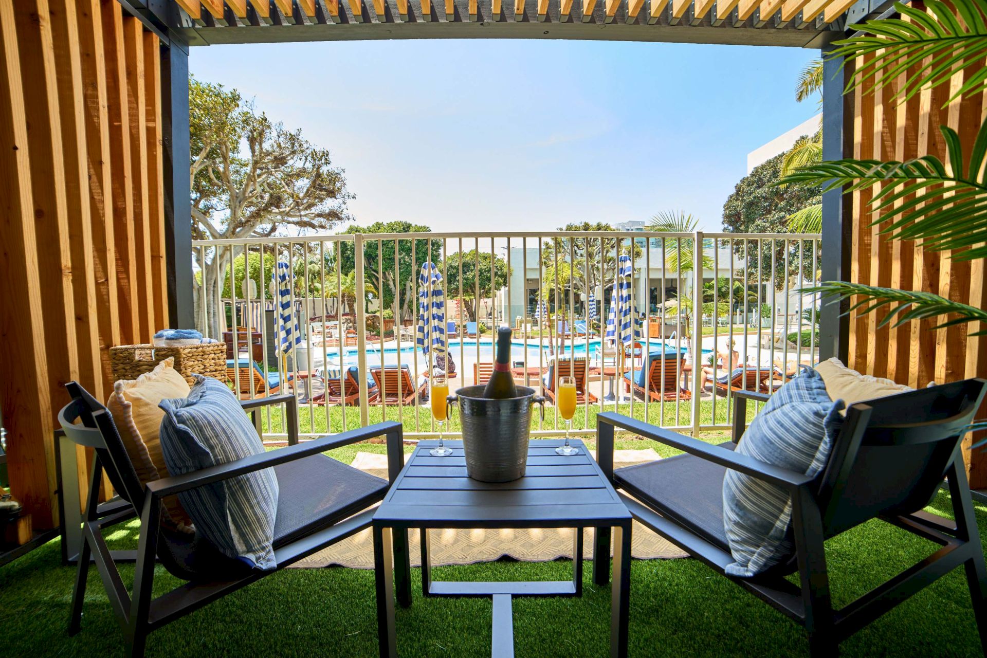 Sunlit ocean-view balcony: cushioned chairs, a table with champagne bucket, and pool umbrellas beyond a white railing, by palm trees.