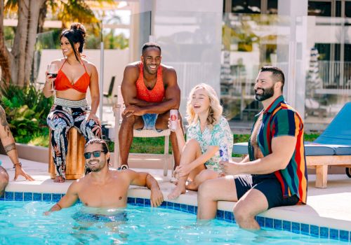 A group of stylish friends relax by a pool: some sit on the edge, one in the water, others stand or chat, enjoying a sunny day together.