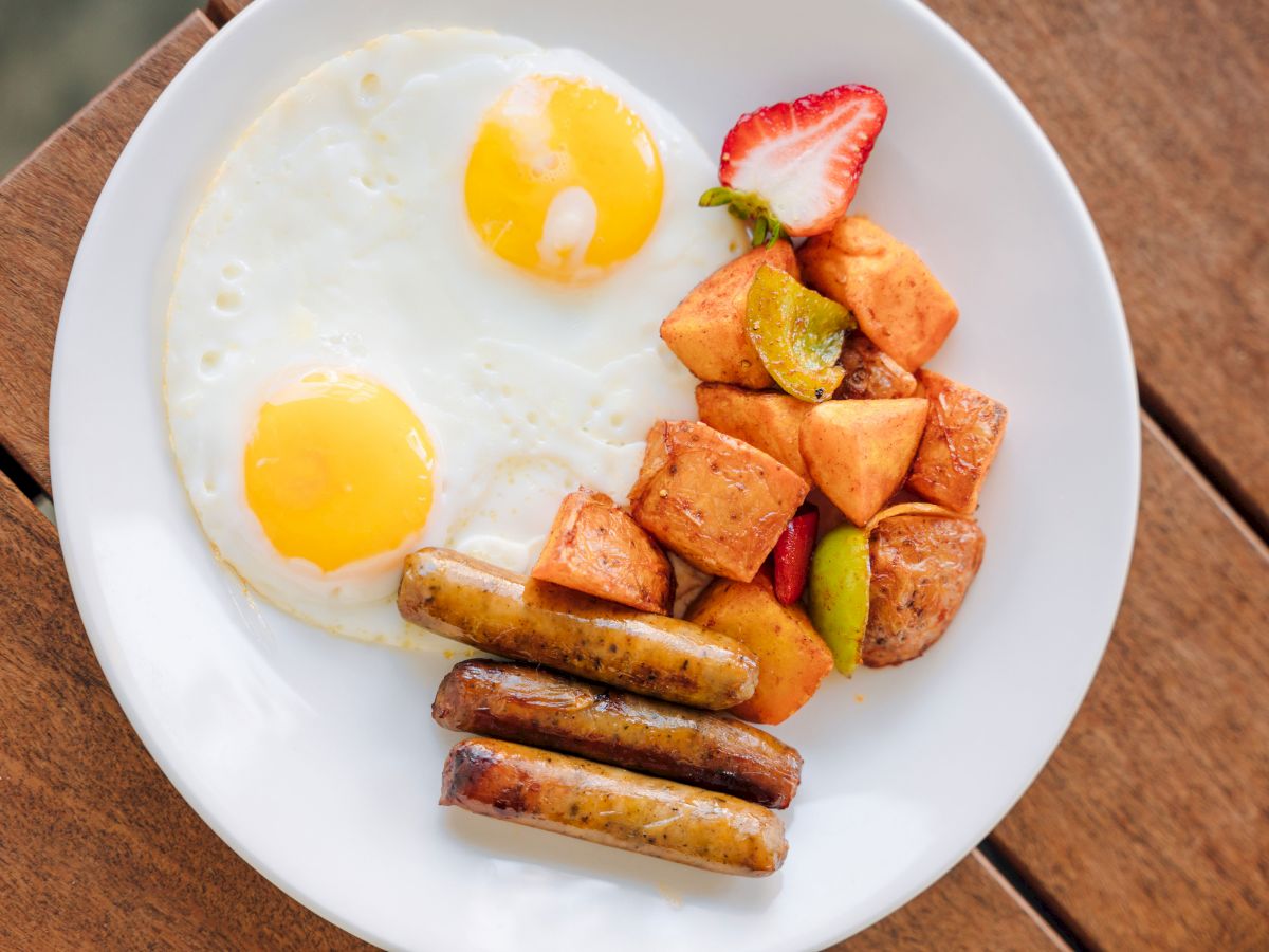 Breakfast plate with two sunny-side eggs, grilled sausages, roasted potatoes/vegetables, and a strawberry garnish.